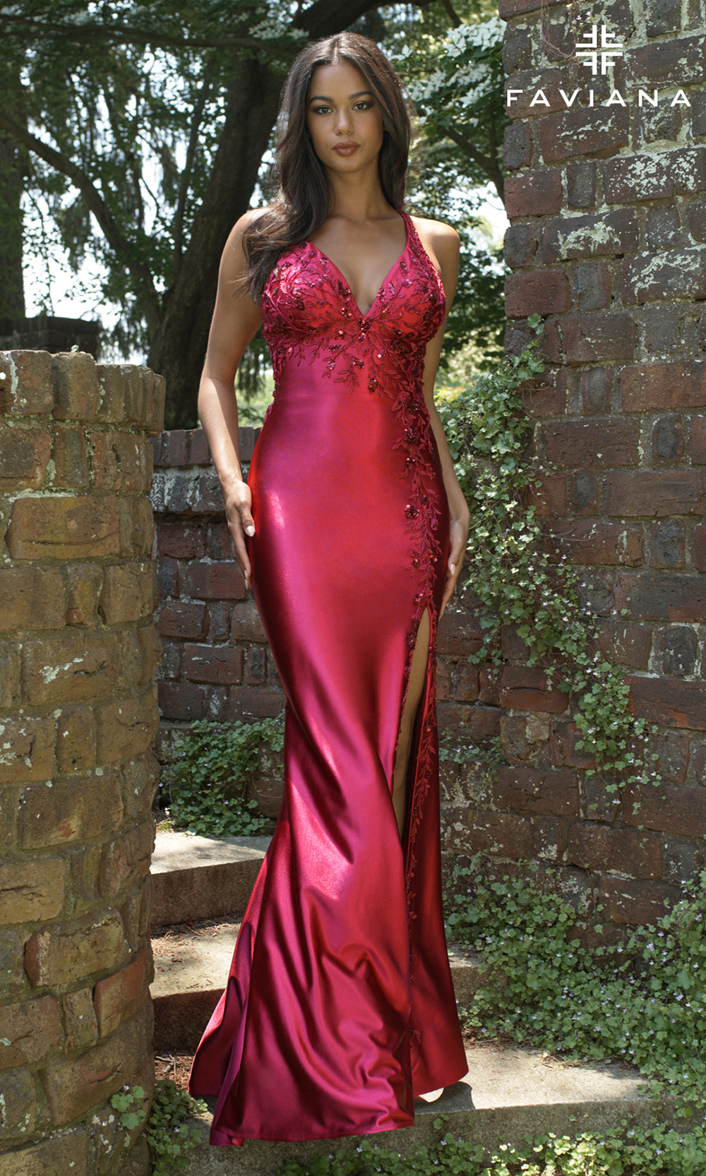 Woman in a red evening gown standing outdoors with a brick wall and greenery in the background.