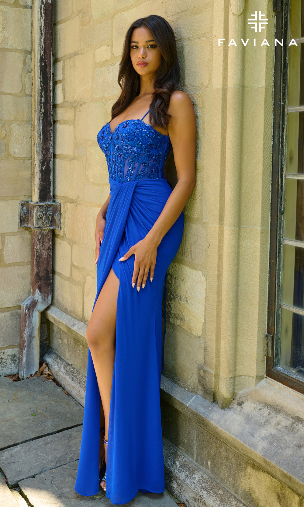 Woman in a blue evening gown standing against a stone wall, with 'Taviana' branding.