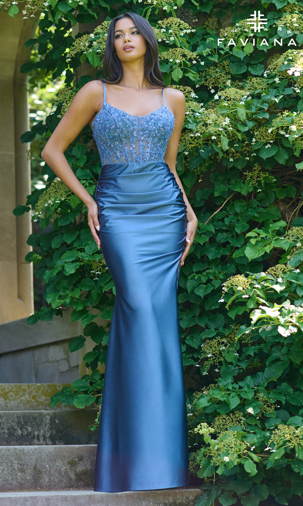 Woman wearing a blue evening gown with sequin top in a garden setting, Favianna brand visible.