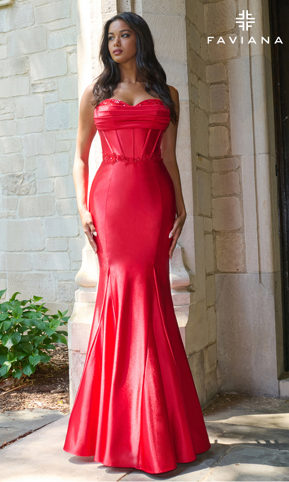 Woman wearing a red evening gown standing against a stone wall, with 'Faviana' branding.