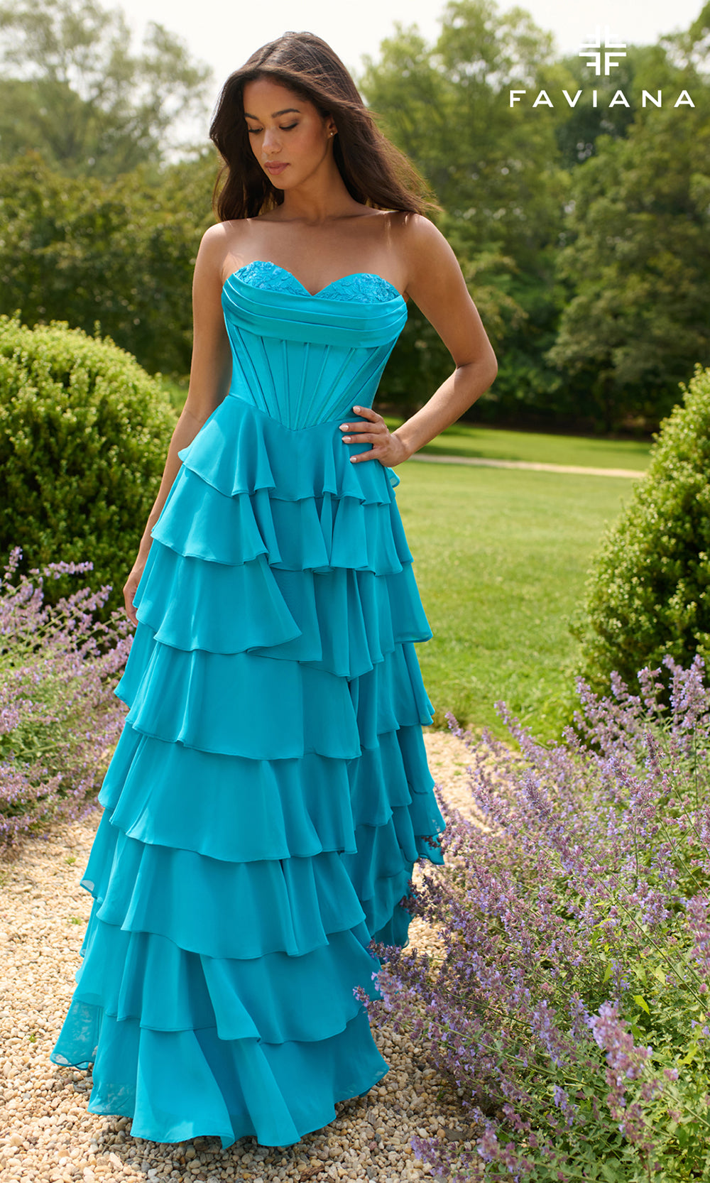 Woman wearing a blue strapless, tiered dress in an outdoor setting with greenery and lavender flowers.