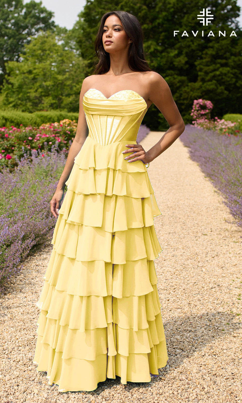 Woman in a yellow strapless gown standing on a path with flowers and trees in the background, FAVIANA brand visible.