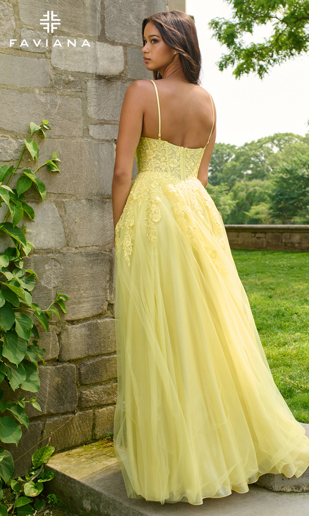 Woman in a yellow dress standing against a stone wall with greenery in the background, FAVIANA brand visible.