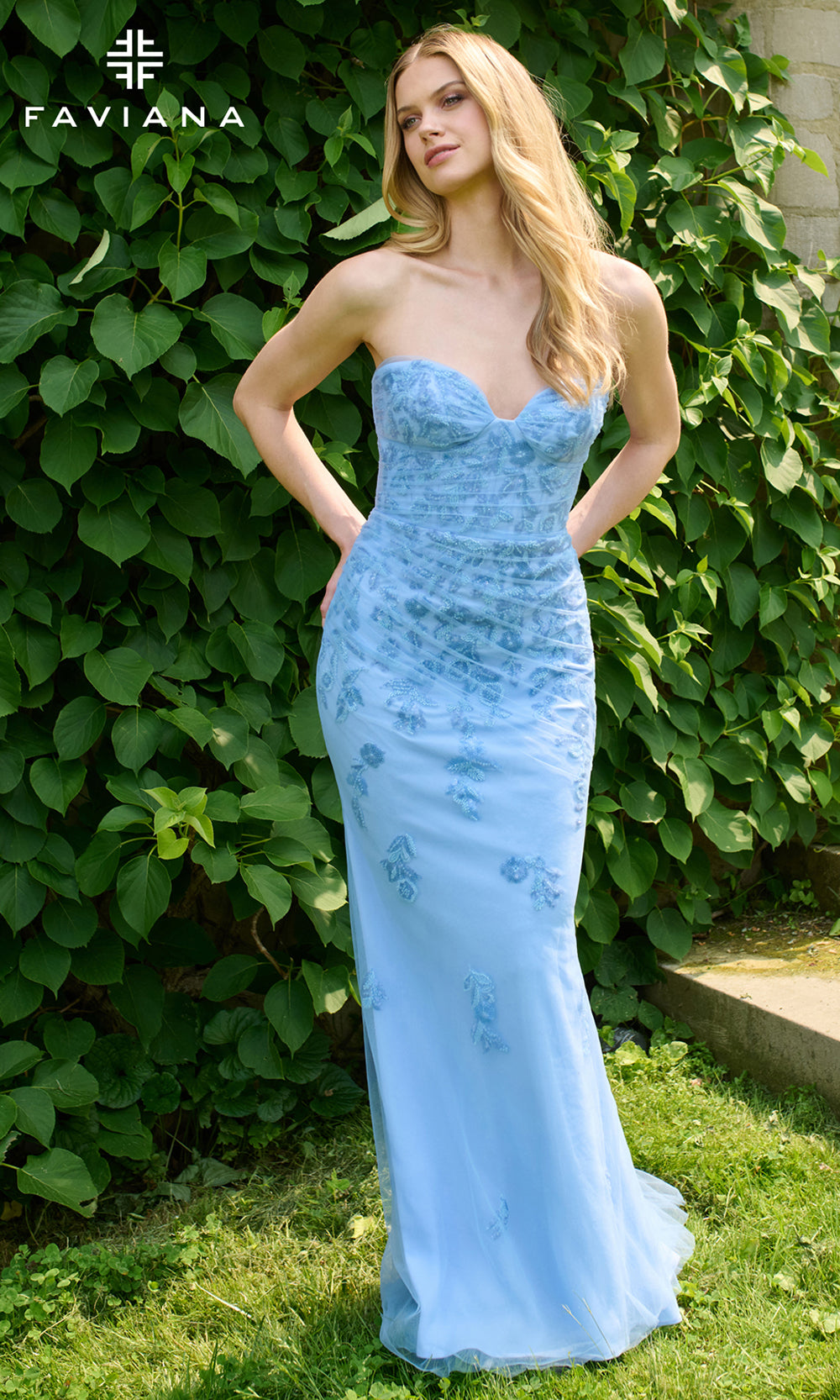 Woman in a light blue strapless gown standing in front of green foliage with FAVIANA branding.