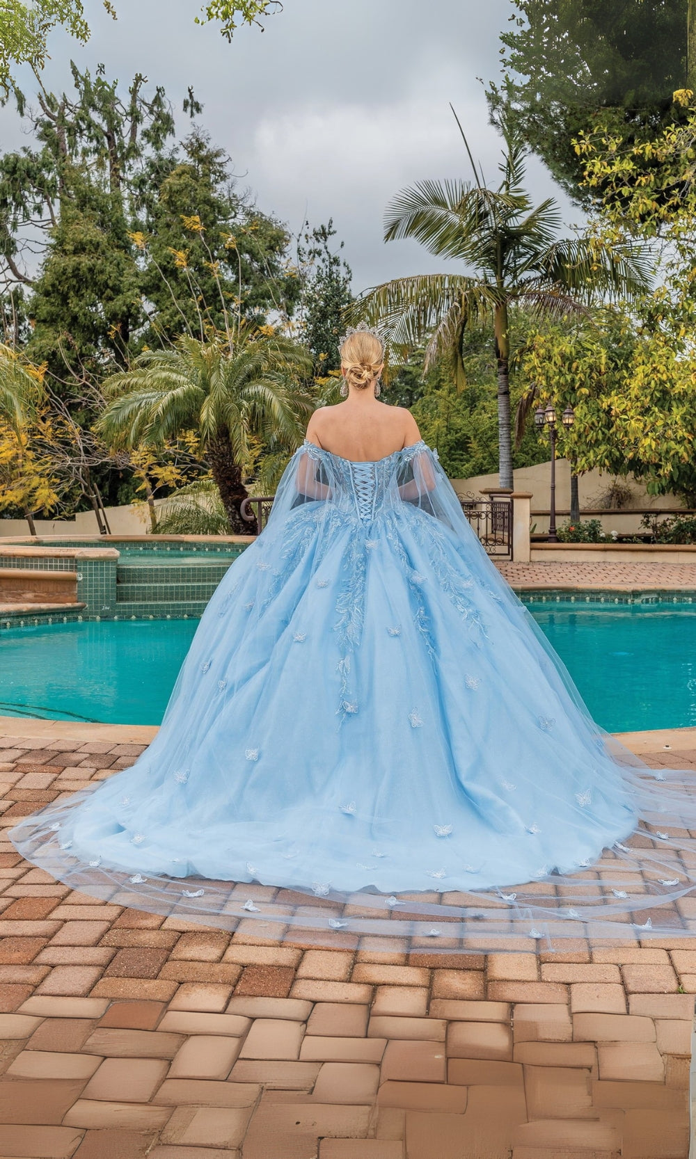 Woman in a light blue ball gown standing by a pool with trees in the background