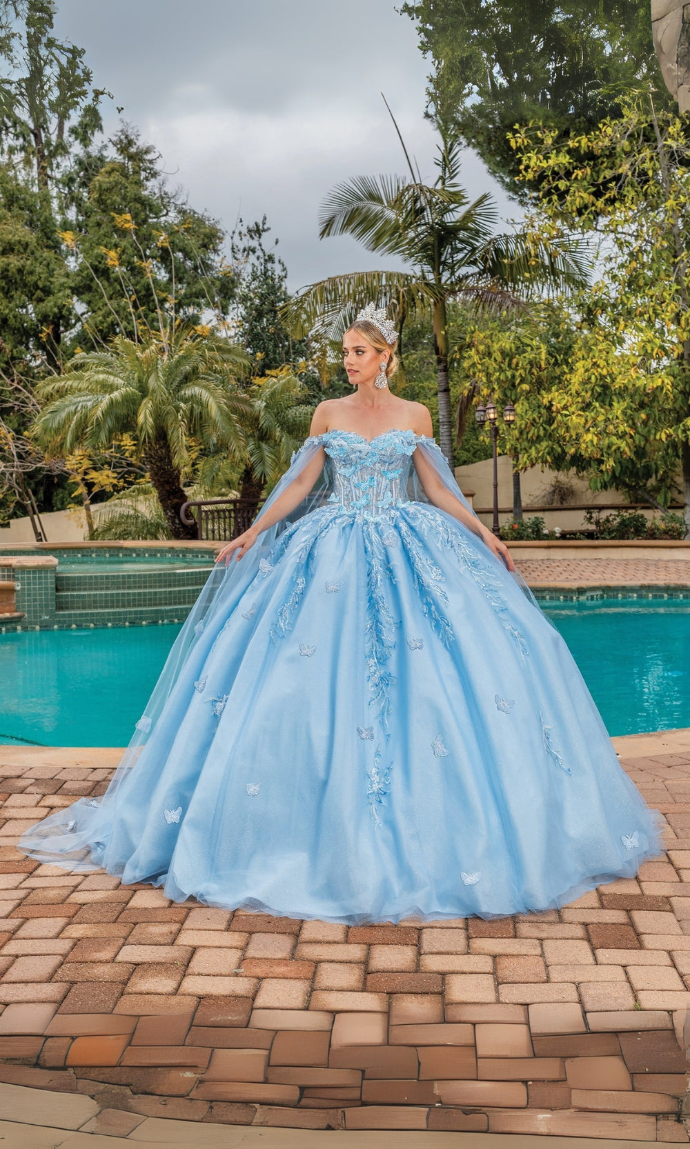 Woman in a light blue ball gown standing by a pool with trees in the background