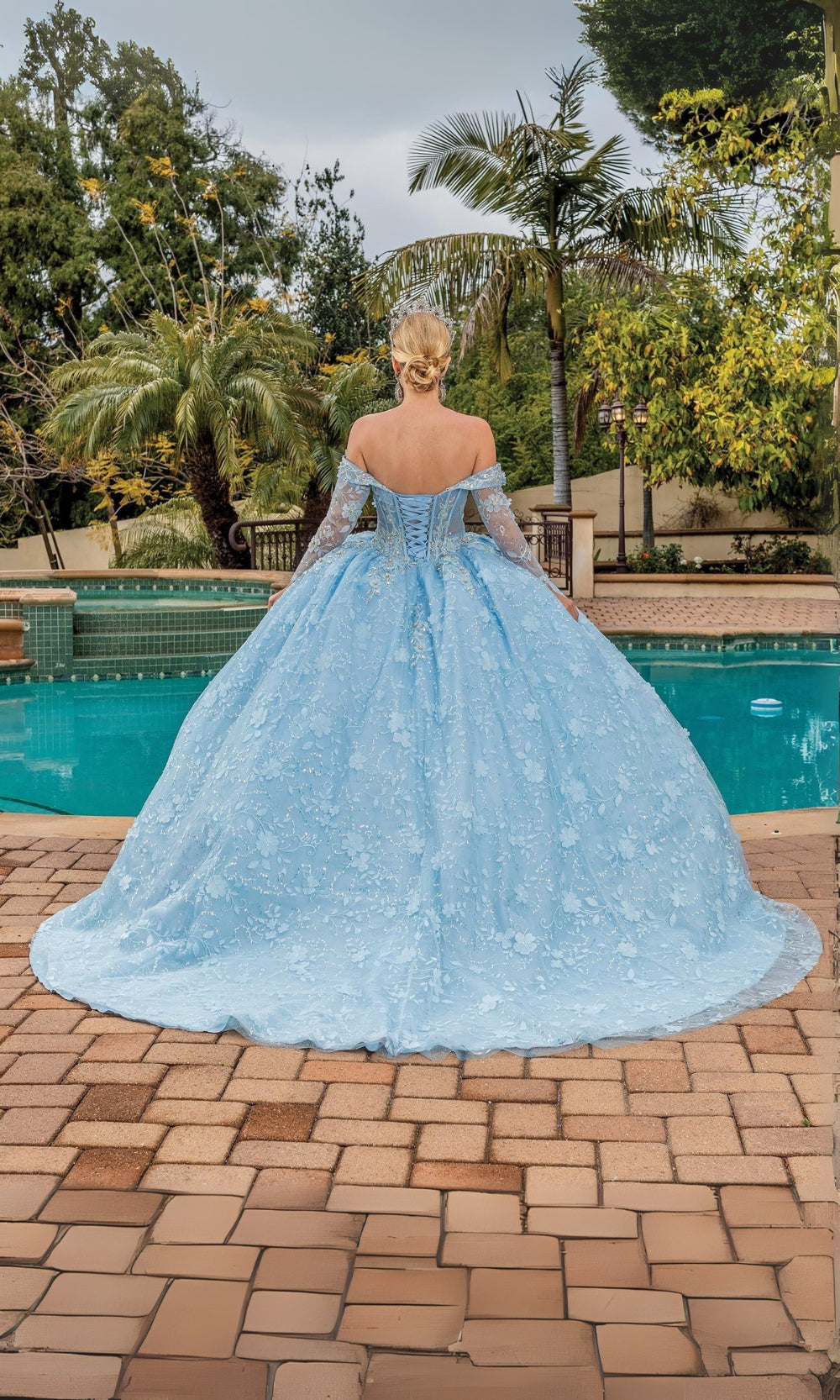 Person wearing a light blue ball gown standing by a pool with trees in the background