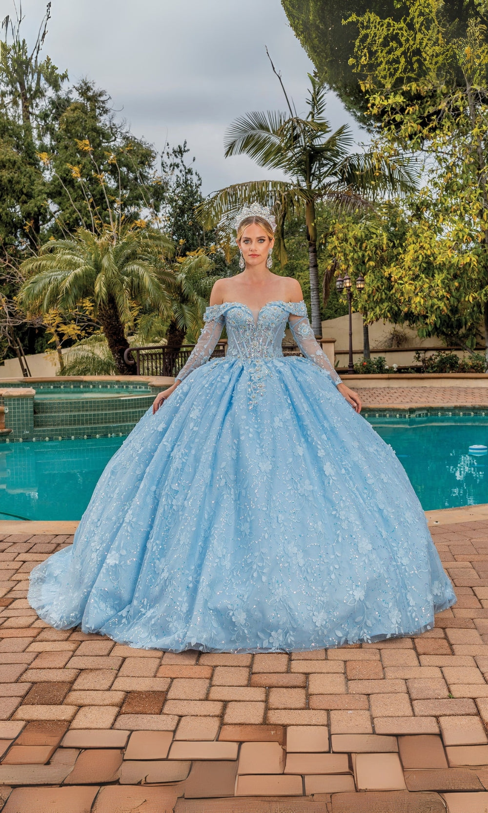 Woman in a blue off-shoulder dress standing by a pool with trees in the background