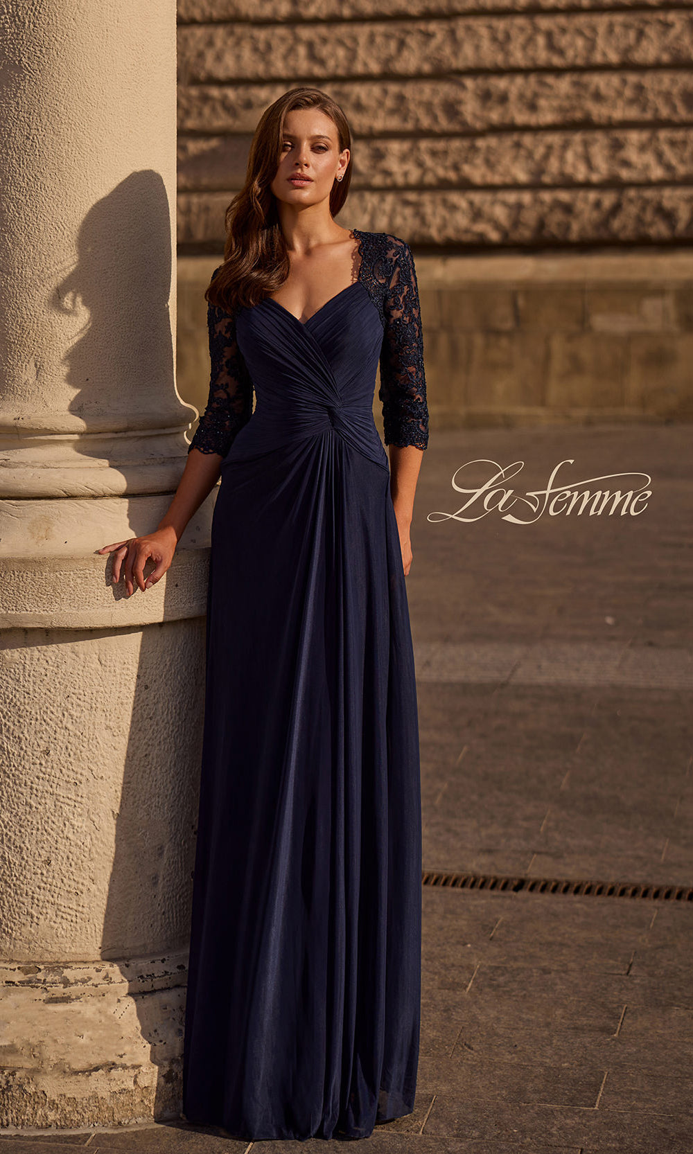 Woman in a navy blue evening gown standing against a stone wall with 'La Femme' branding.