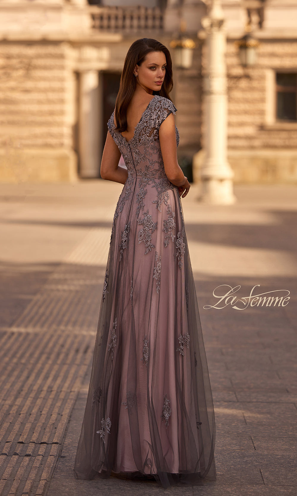 Woman in a long, elegant dress with floral embellishments standing on a stone pavement.