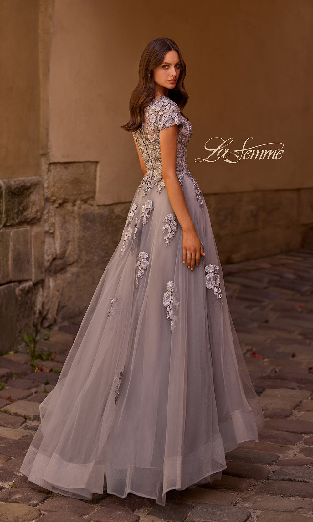 Woman in a light purple gown with floral embellishments standing against a stone wall.