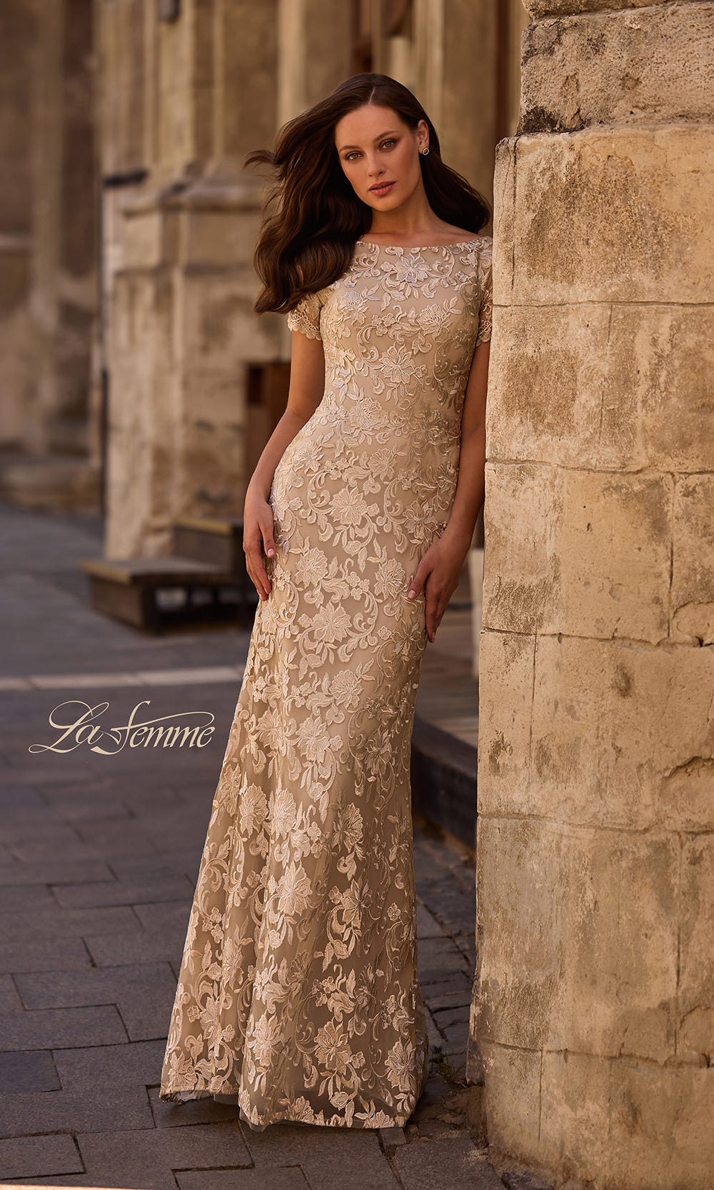 Woman in a gold lace dress standing against a stone wall.