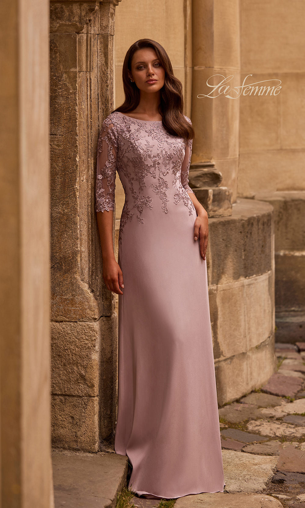 Woman in a long, light pink dress with lace detailing standing against a stone wall.