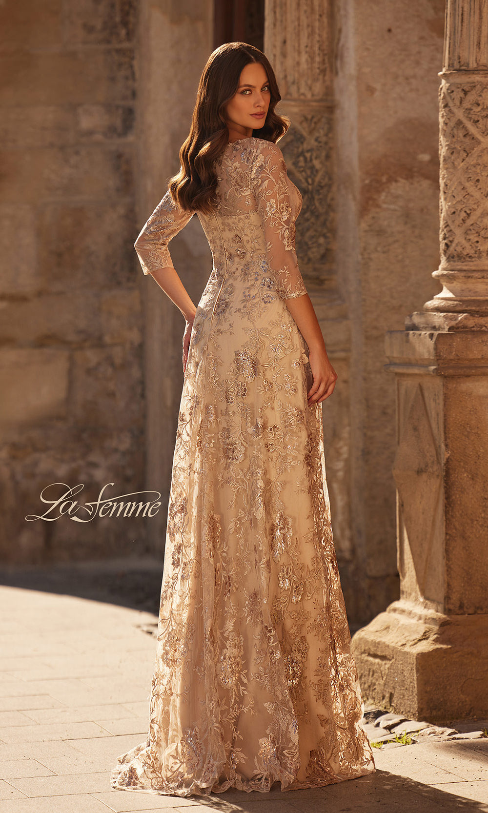 Woman in a beige lace evening gown standing in front of stone architectural elements.