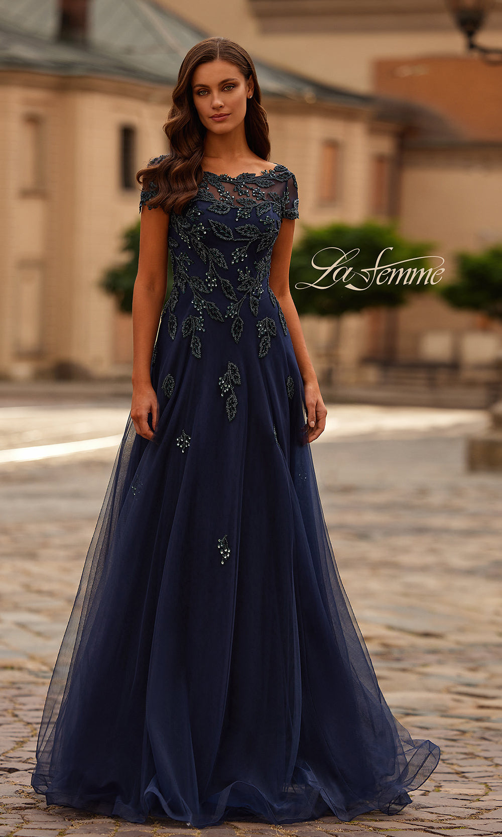 Woman wearing a dark blue evening gown with intricate lace details on a blurred outdoor background.