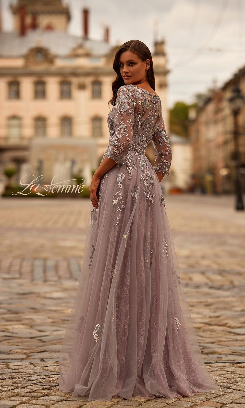 Woman in a pink evening gown with floral embellishments standing on a cobblestone street.