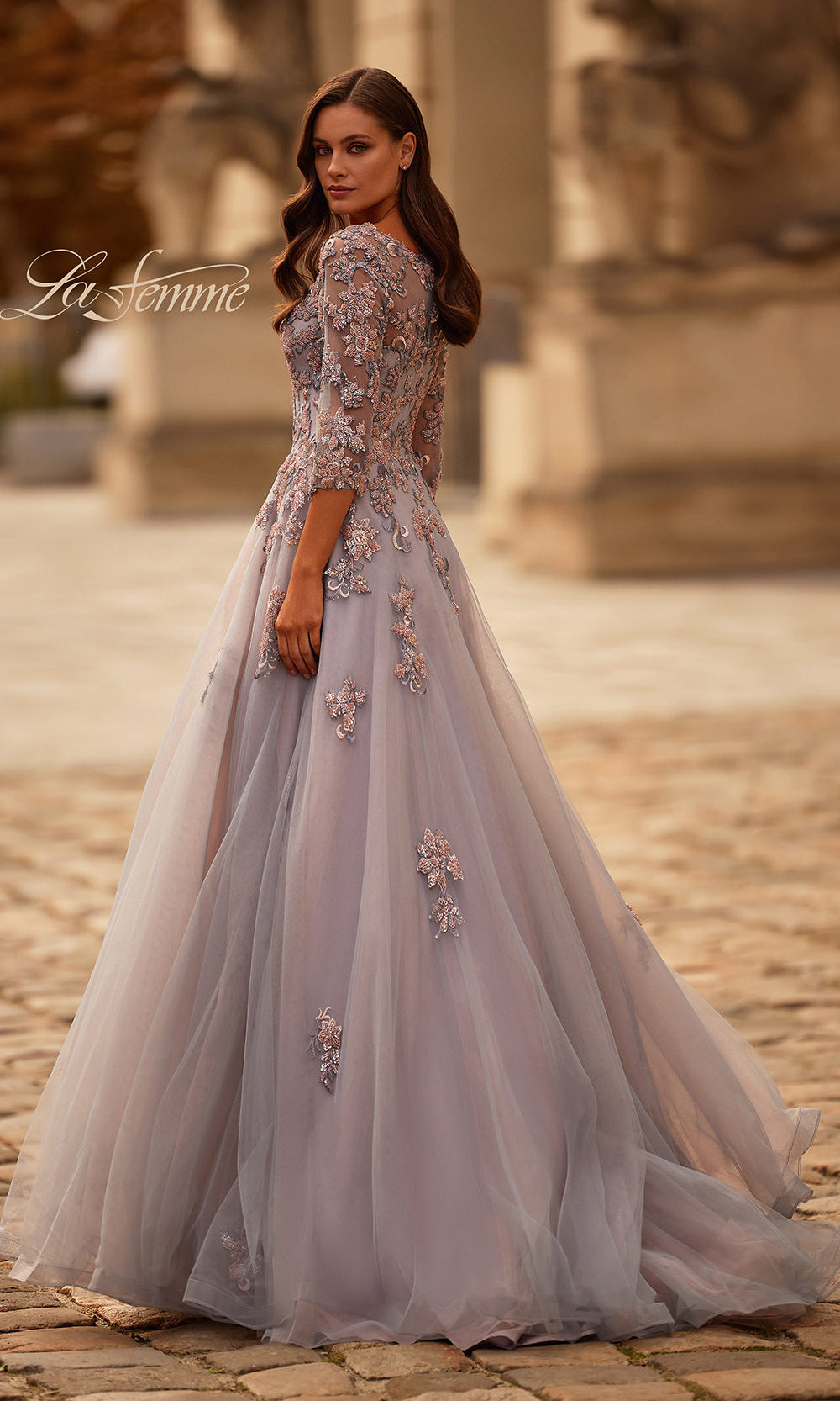 Woman in an elegant evening gown with floral details standing in a historic setting.