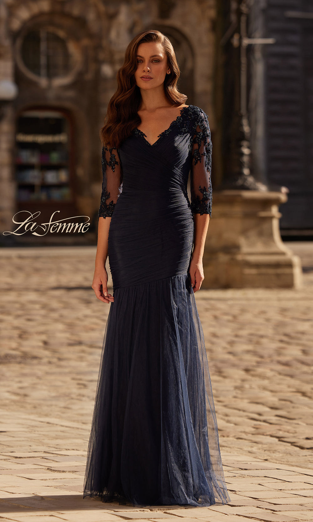 Woman wearing a dark blue evening gown with sheer sleeves on a cobblestone street.