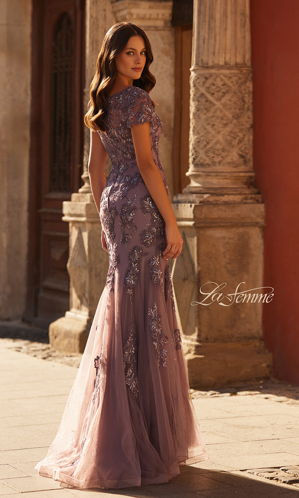 Woman in a purple evening gown standing in an outdoor setting with architectural elements.