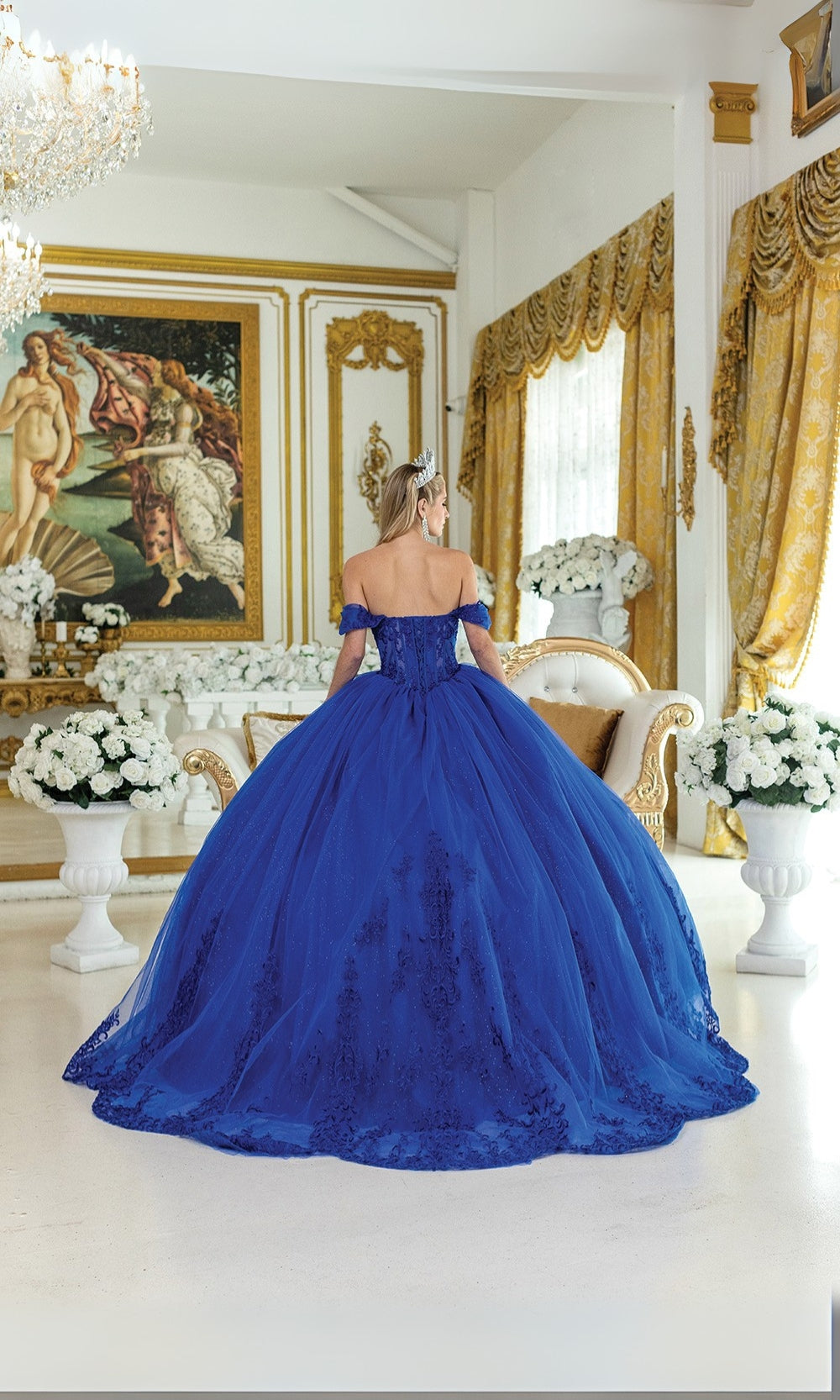 Woman in a blue ball gown standing in an ornate room with gold accents and white flowers.