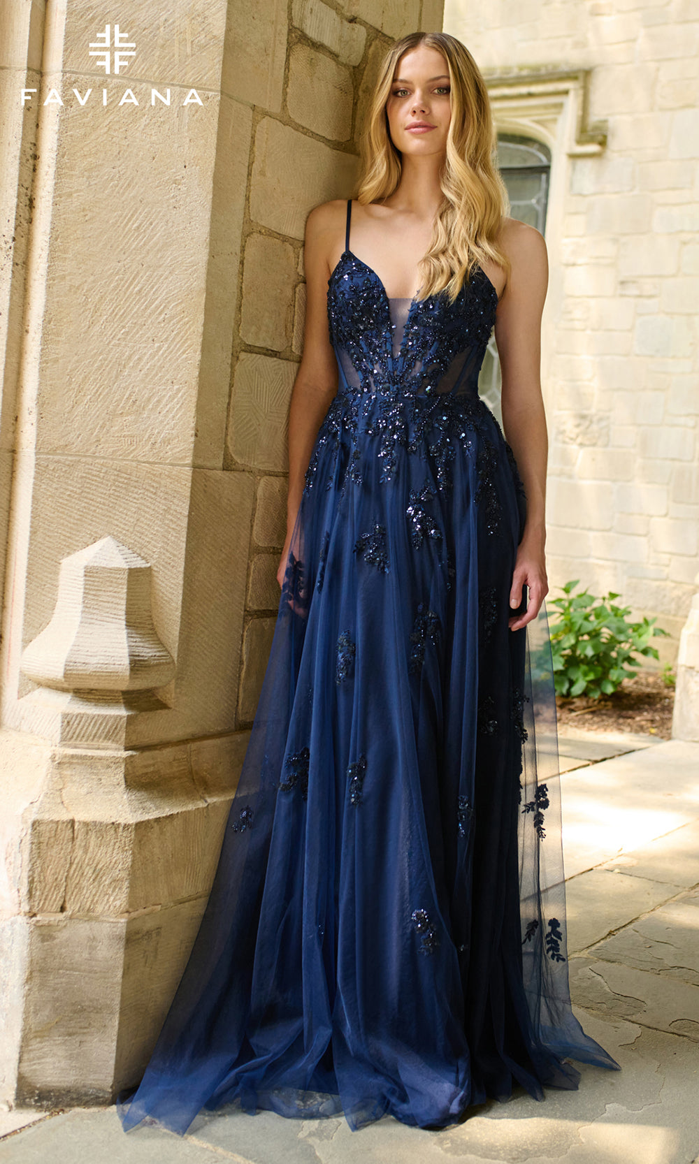 Woman wearing a blue evening gown with floral embellishments against a stone wall.