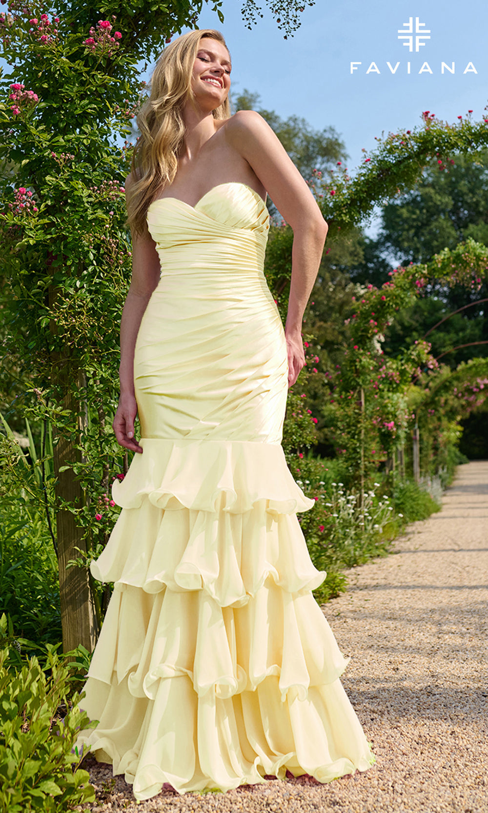 Woman in a light yellow strapless gown with ruffled layers standing in a garden setting, with 'Faviana' branding.