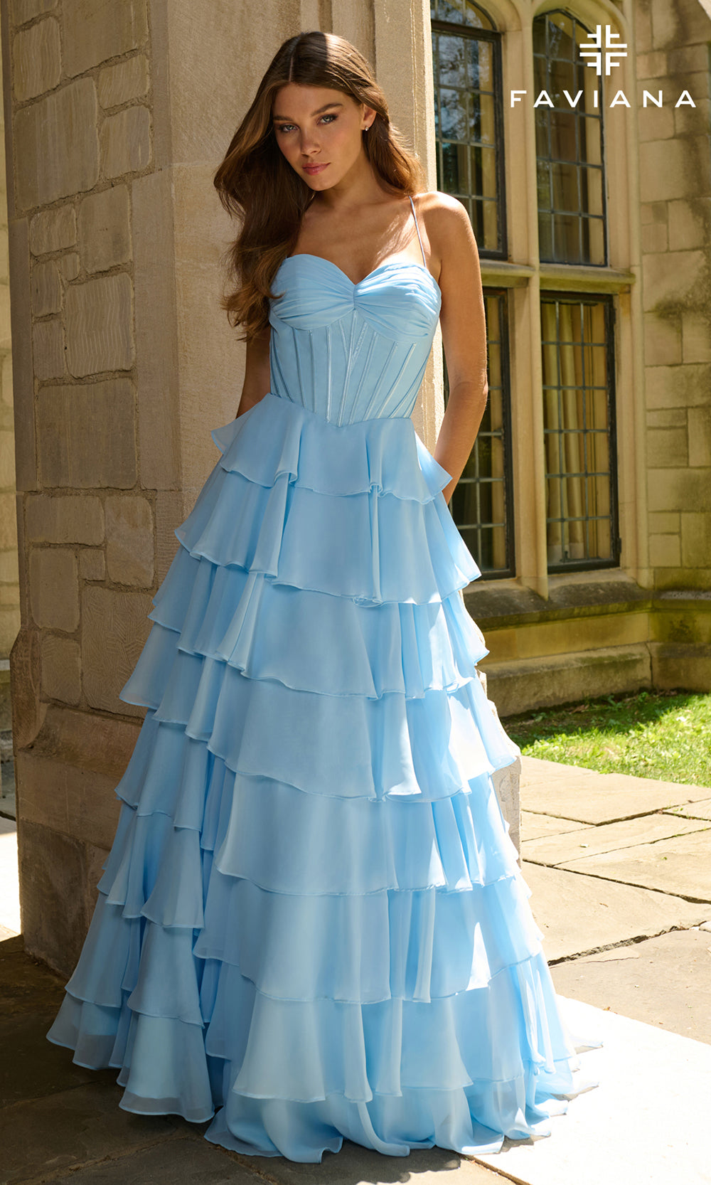 Woman wearing a light blue tiered dress in front of a stone building with 'Faviana' branding.
