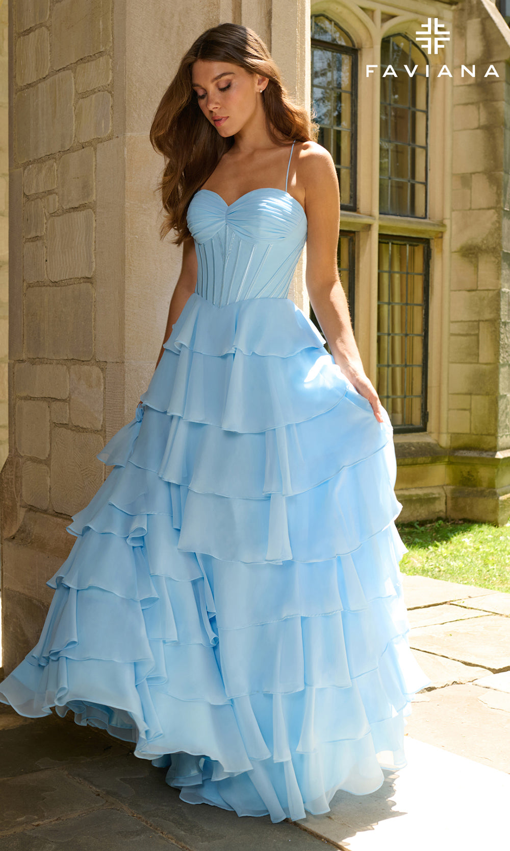 Woman wearing a light blue ruffled dress in front of a stone building with 'Taviana' branding.