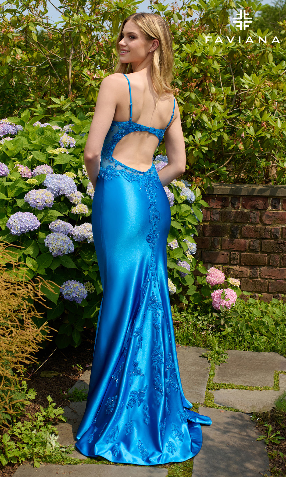 Woman in a blue evening gown standing in a garden with flowers and a brick wall.