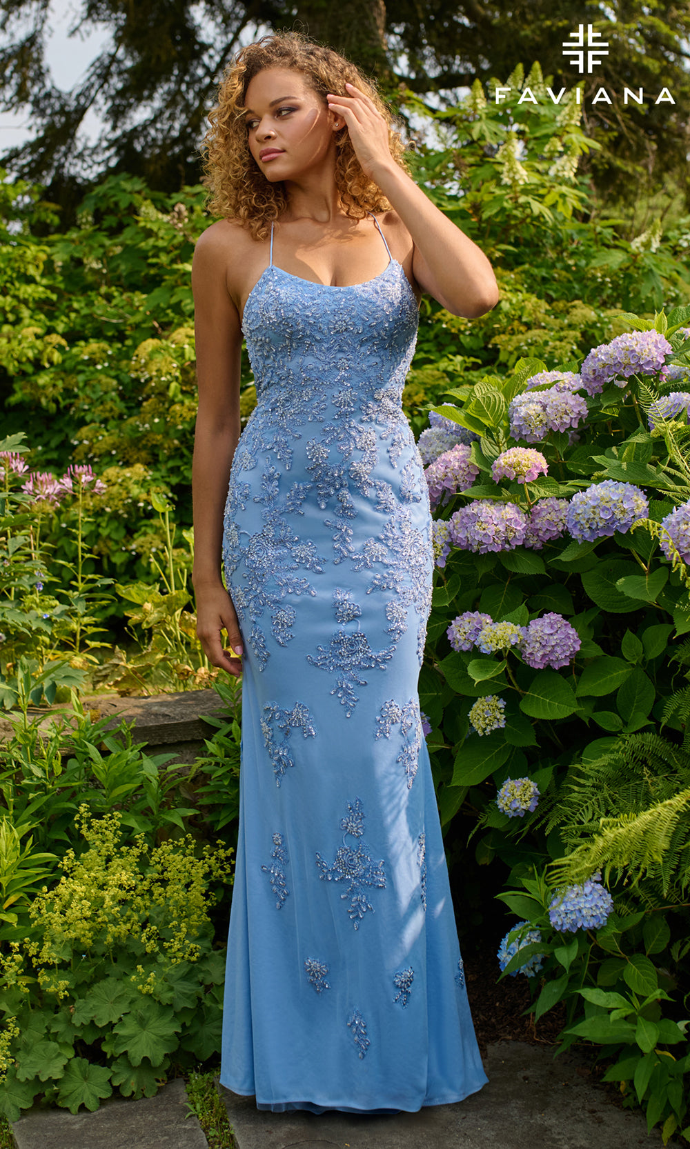 Woman in a blue evening gown standing in a garden with flowers and trees.