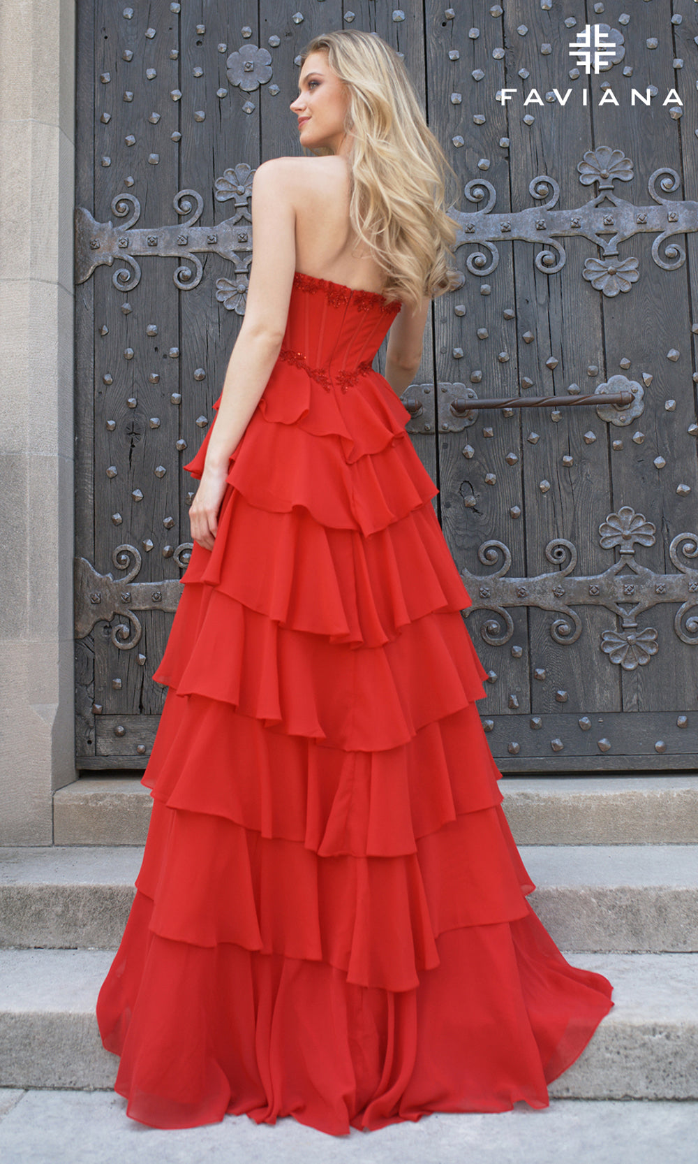 Woman in a red strapless, tiered gown standing in front of an ornate door with 'Faviana' branding.