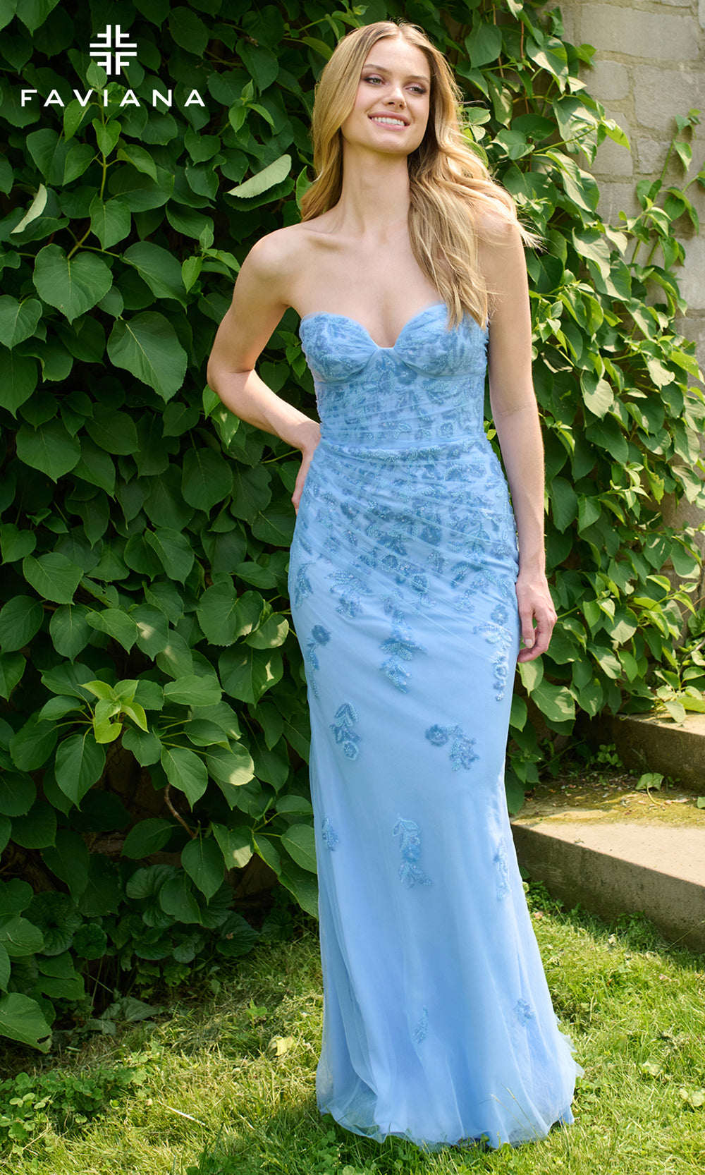 Woman wearing a light blue strapless gown with floral patterns in an outdoor setting, with FAVIANA branding.