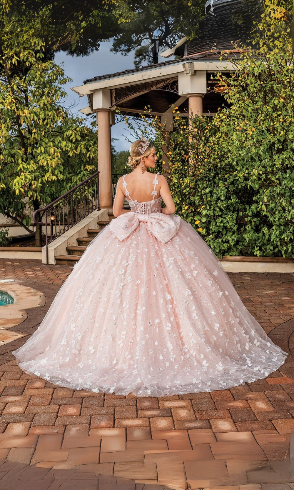 Woman in a pink ball gown with a large bow standing outdoors on a brick patio.
