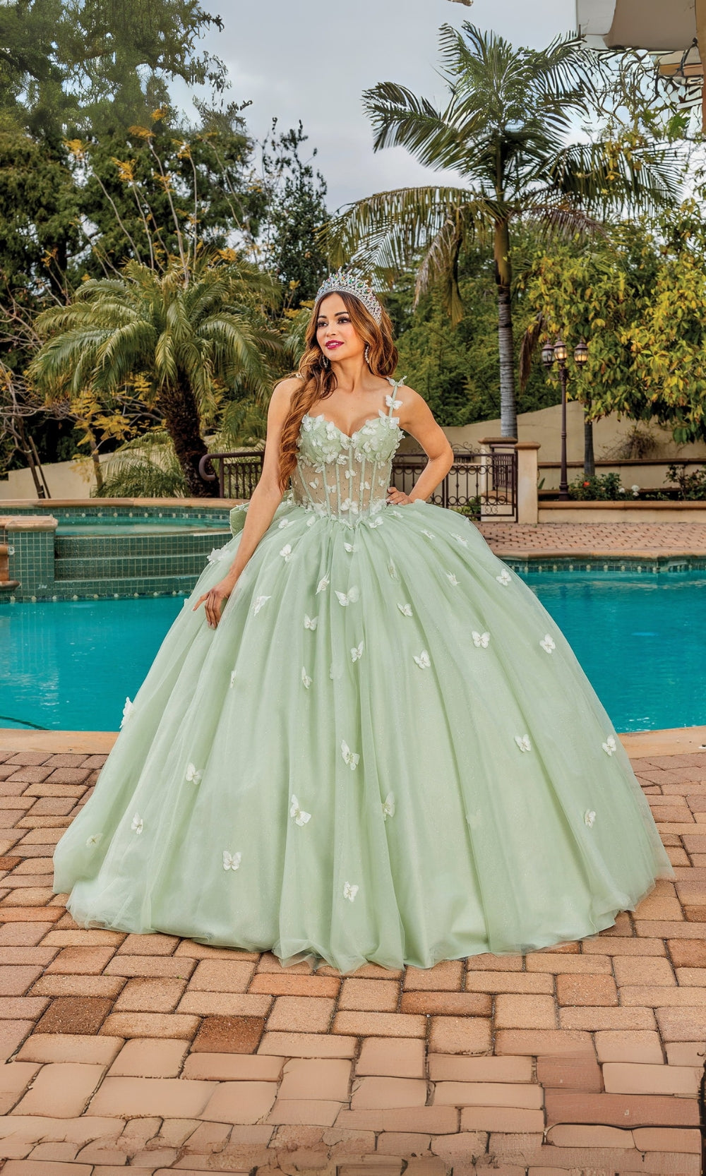 Woman in a light green ball gown standing by a pool with palm trees in the background