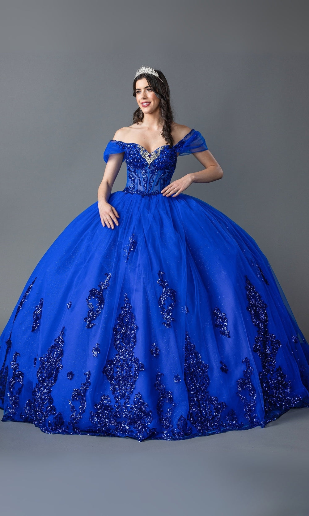 Woman wearing a blue off-shoulder gown with intricate detailing on a gray background