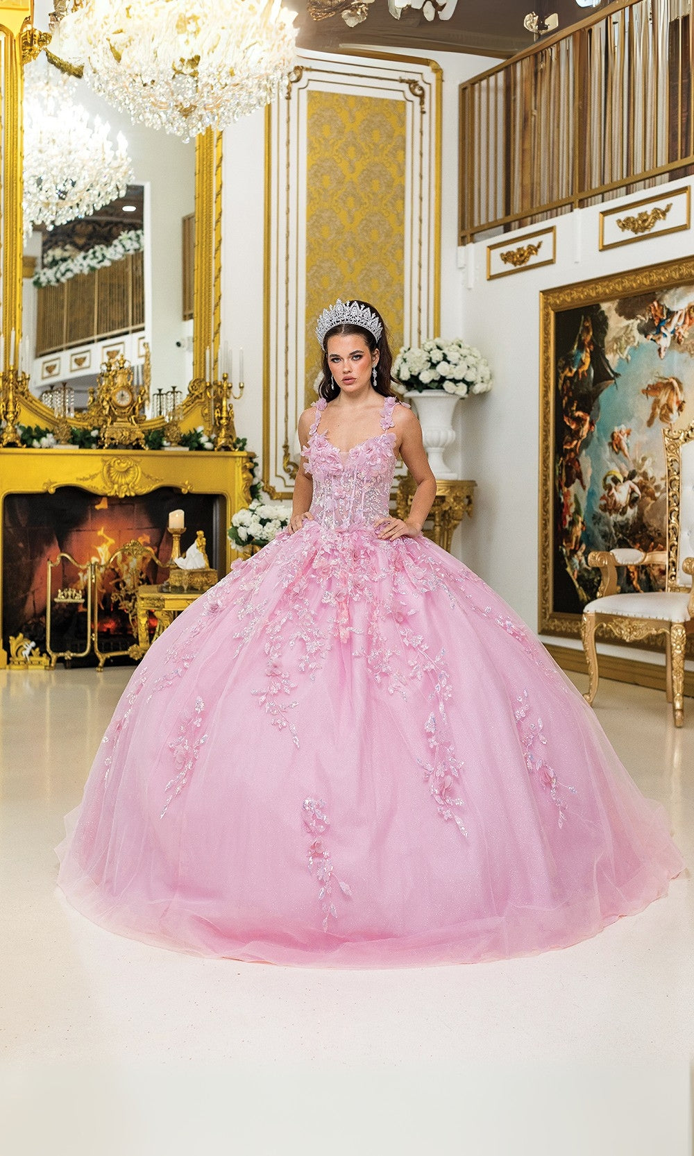 Woman in a pink gown standing in an ornate room with chandelier and decorative wall.
