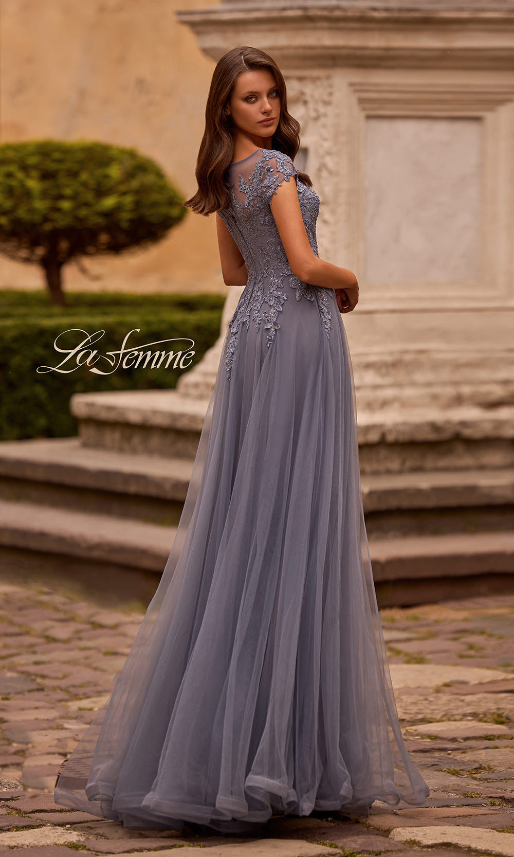 Woman in a gray evening gown standing in an outdoor setting with classical architecture.