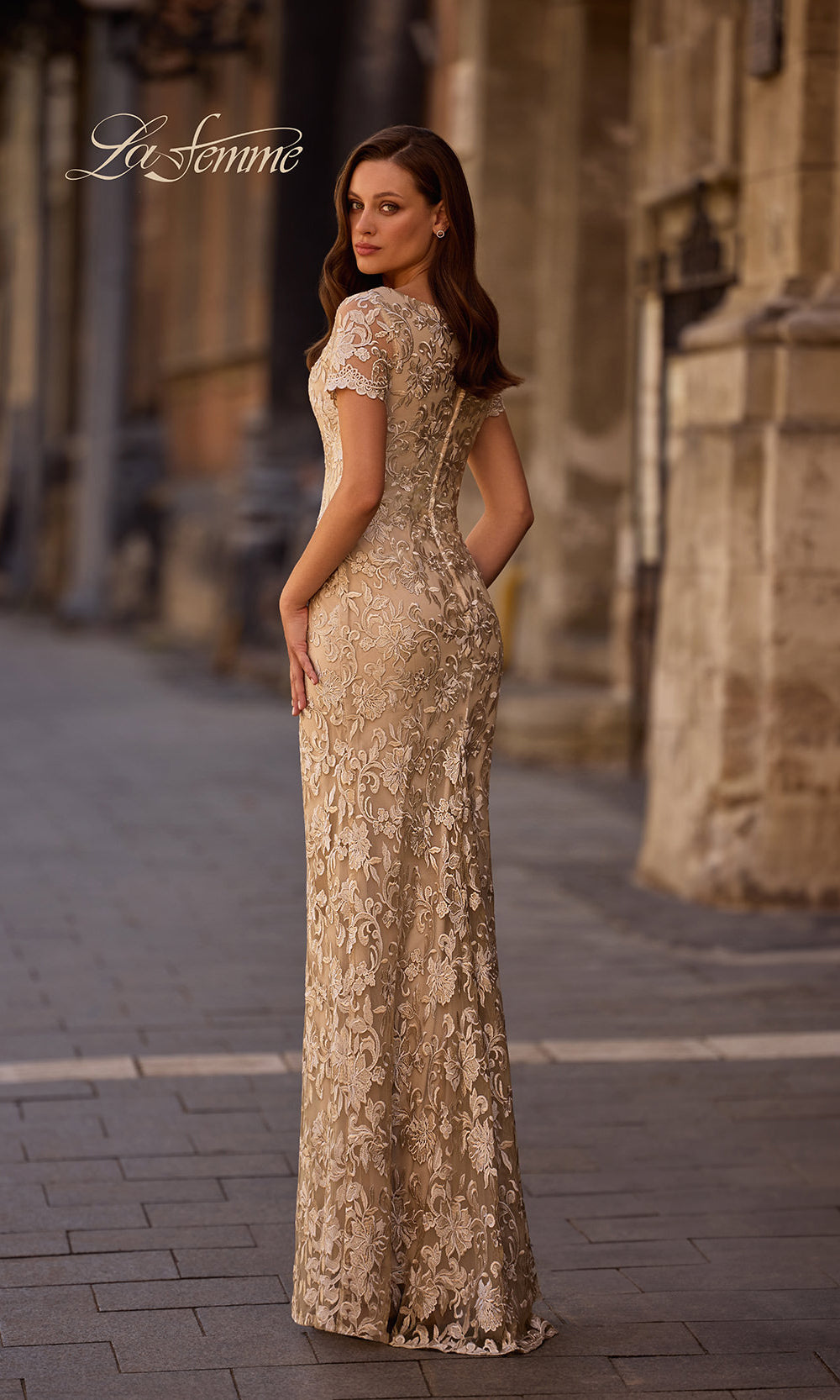 Woman in a long, elegant dress standing on a street with architectural columns in the background.