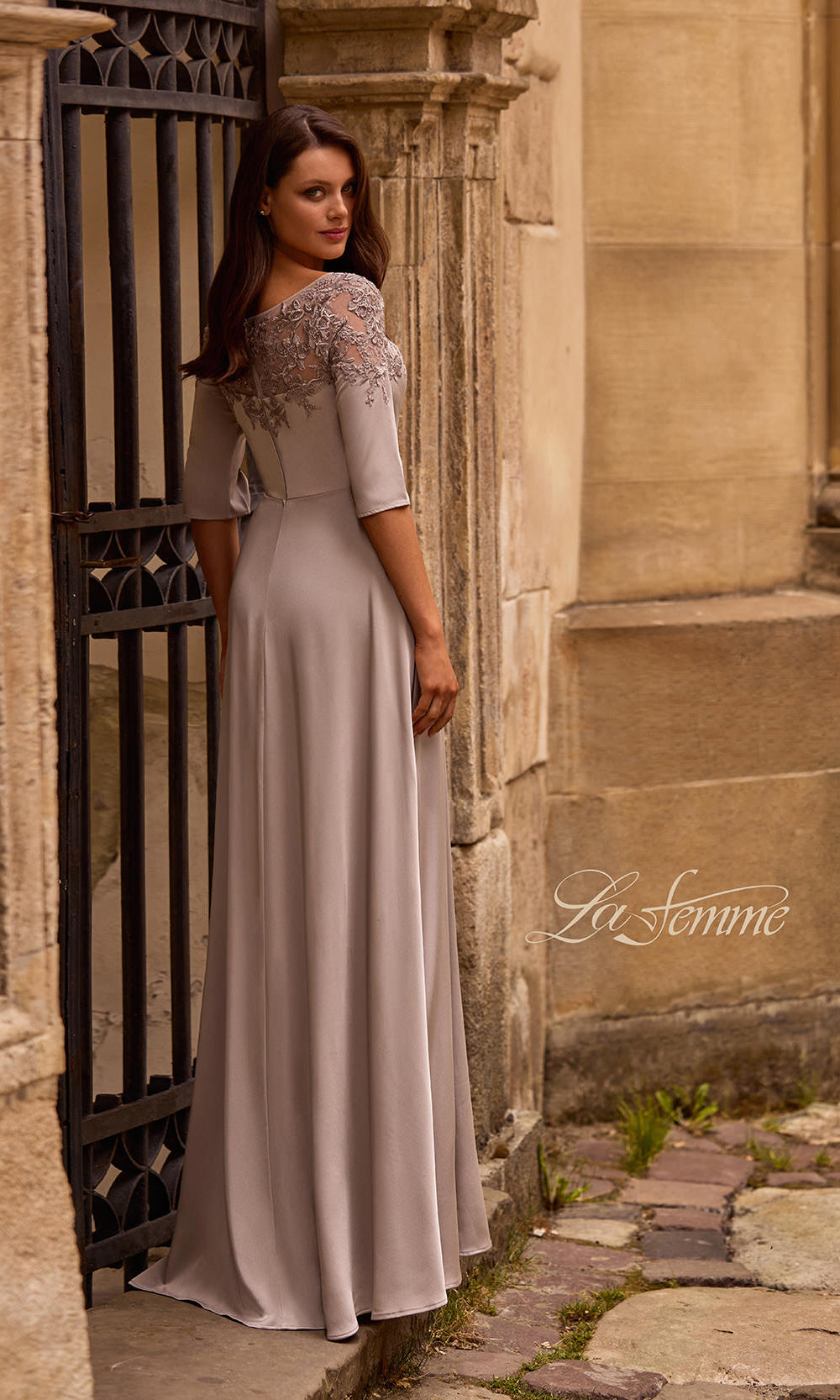 Woman in a long, elegant dress standing in an outdoor setting with stone walls and a gate.