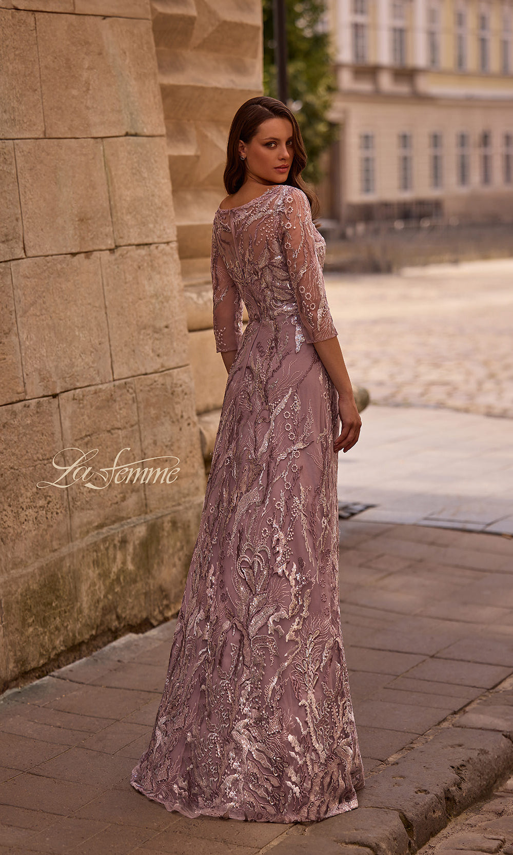 Woman in a long, elegant dress standing against a stone wall with a blurred building in the background.