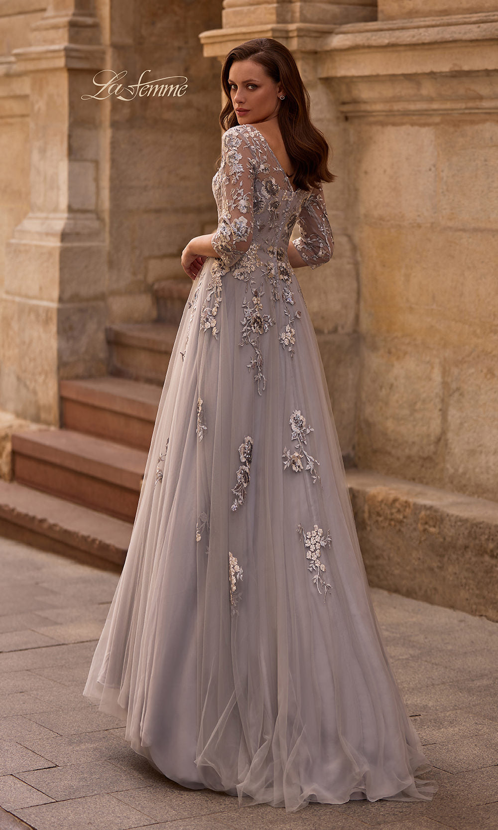 Woman in a silver evening gown with floral embellishments standing in front of stone steps.