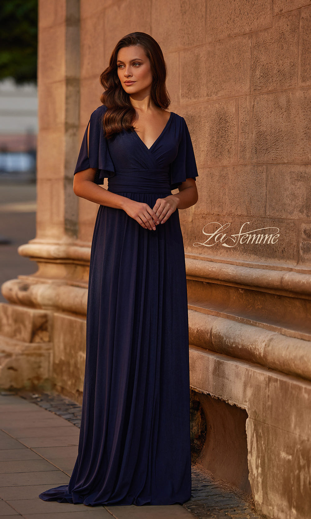 Woman in a navy blue dress standing against a stone wall with 'La Femme' branding.
