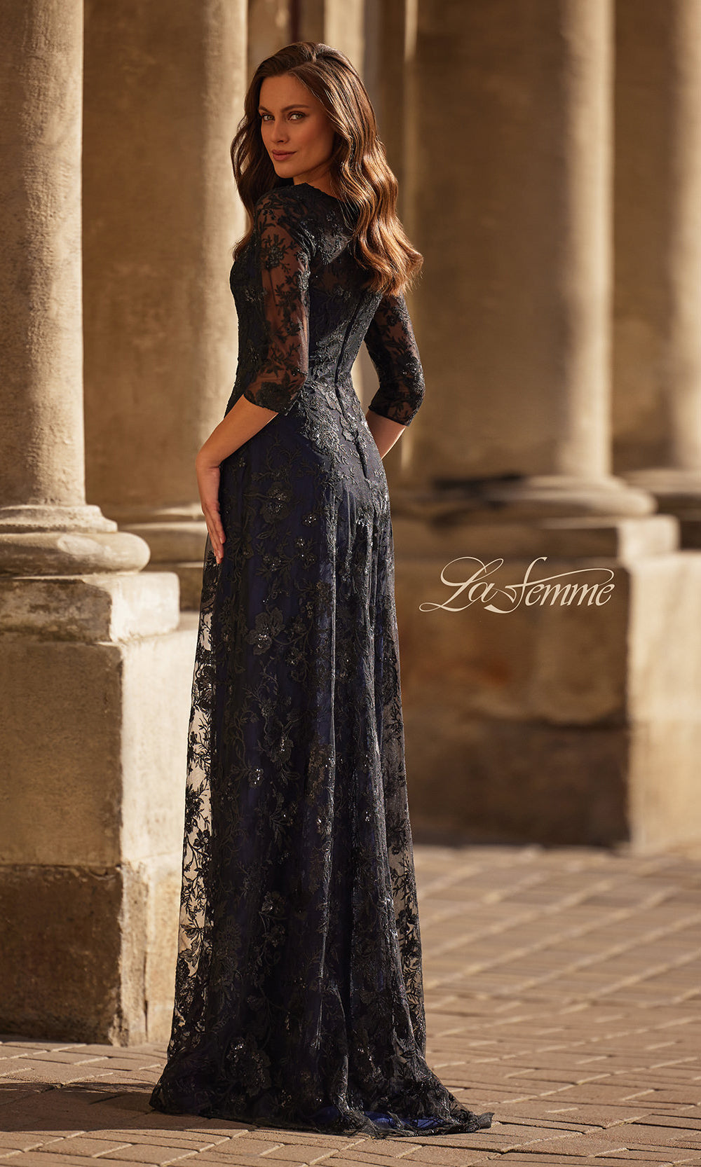 Woman in a long, dark blue lace dress standing in front of classical architecture.