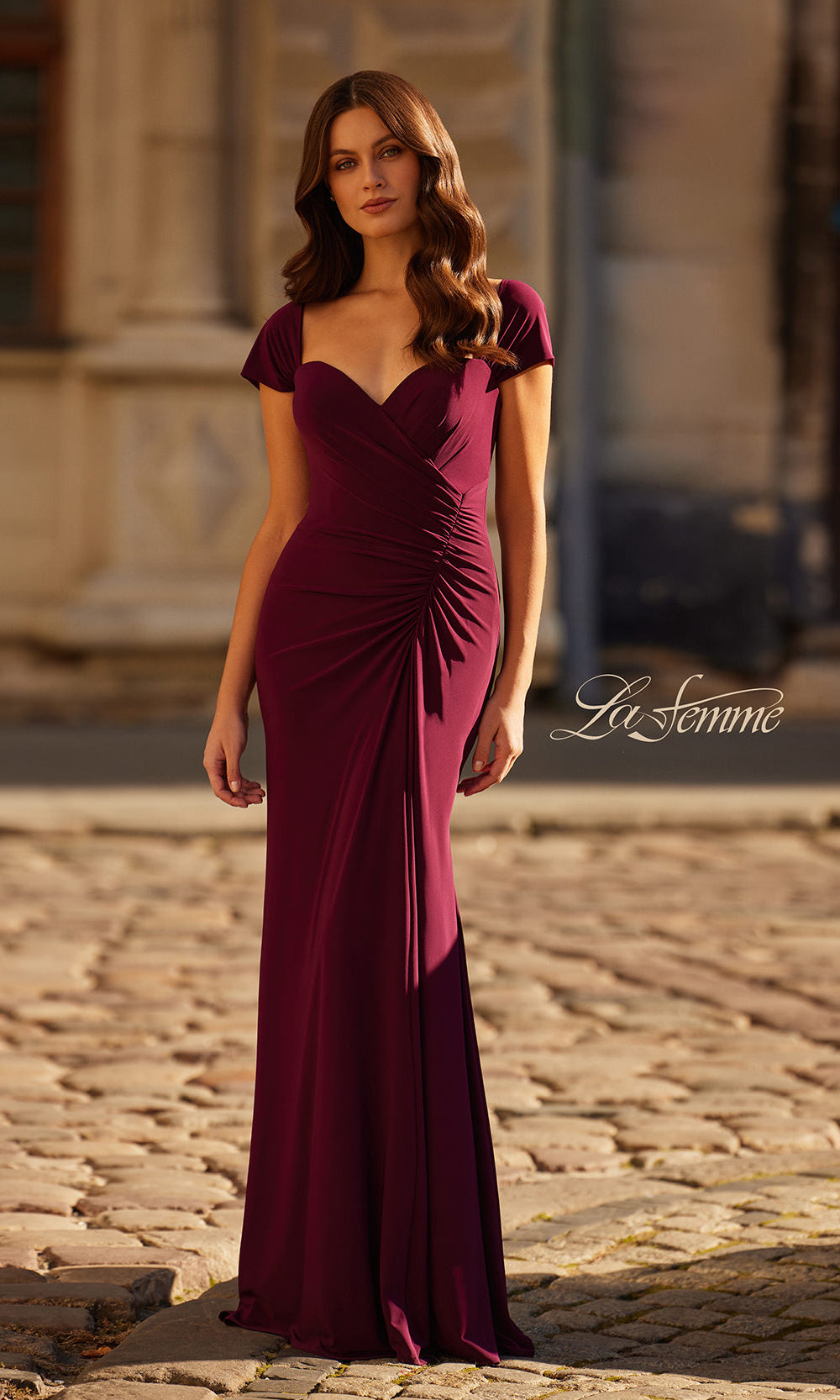 Woman wearing a burgundy evening gown on a cobblestone street with 'La Femme' branding.