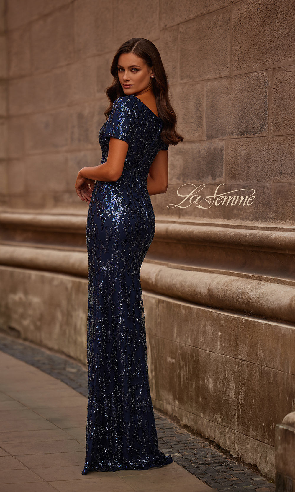 Woman in a long blue sequin dress standing against a stone wall with 'La Femme' branding.