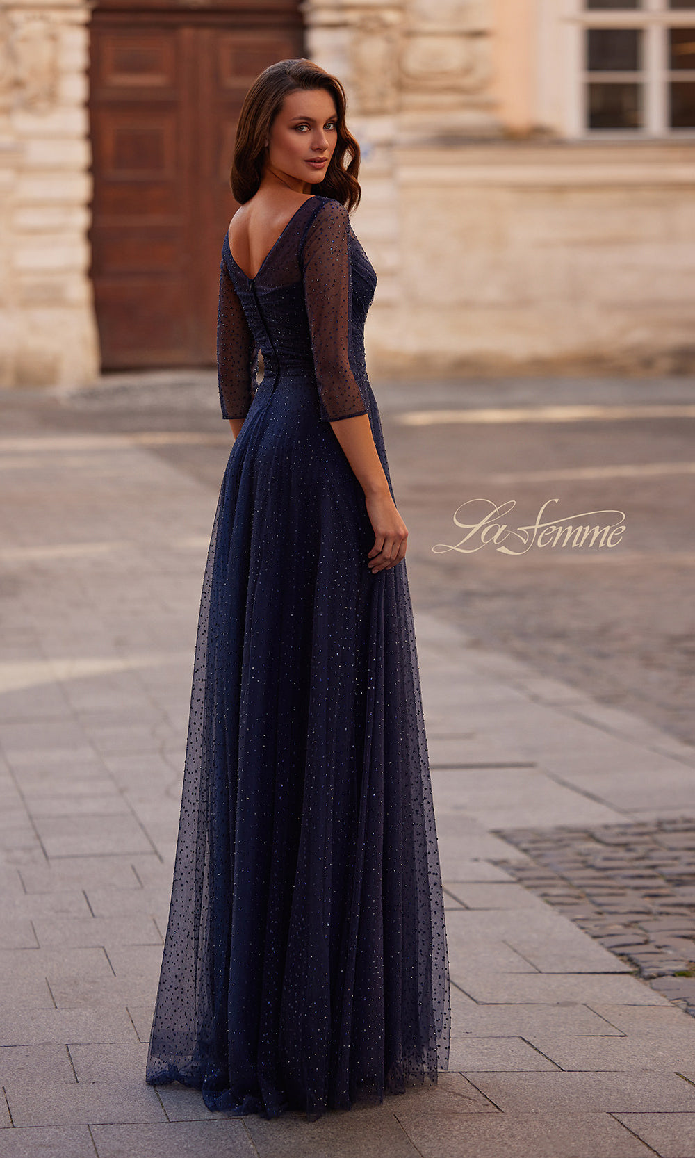 Woman wearing a dark blue evening gown with a sheer overlay in an outdoor setting.
