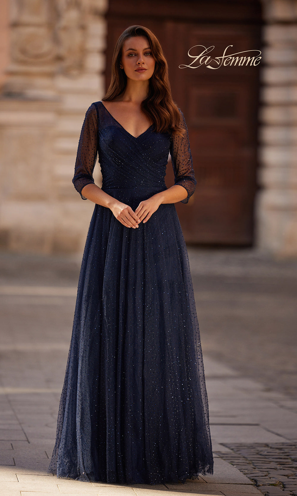 Woman wearing a dark blue evening gown with sheer sleeves in an outdoor setting.