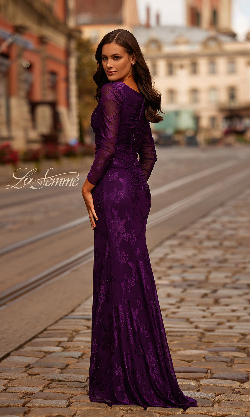 Woman in a purple evening gown standing on a cobblestone street with blurred buildings in the background.