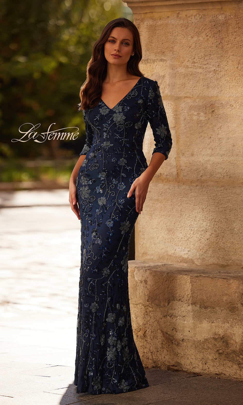 Woman wearing a navy blue floral evening gown standing against a stone wall.